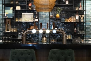 A bar area with a black brick wall, shelves with bottles and glasses, and two green upholstered chairs. - Hotel De Molenhoek - Nijmegen (Molenhoek)