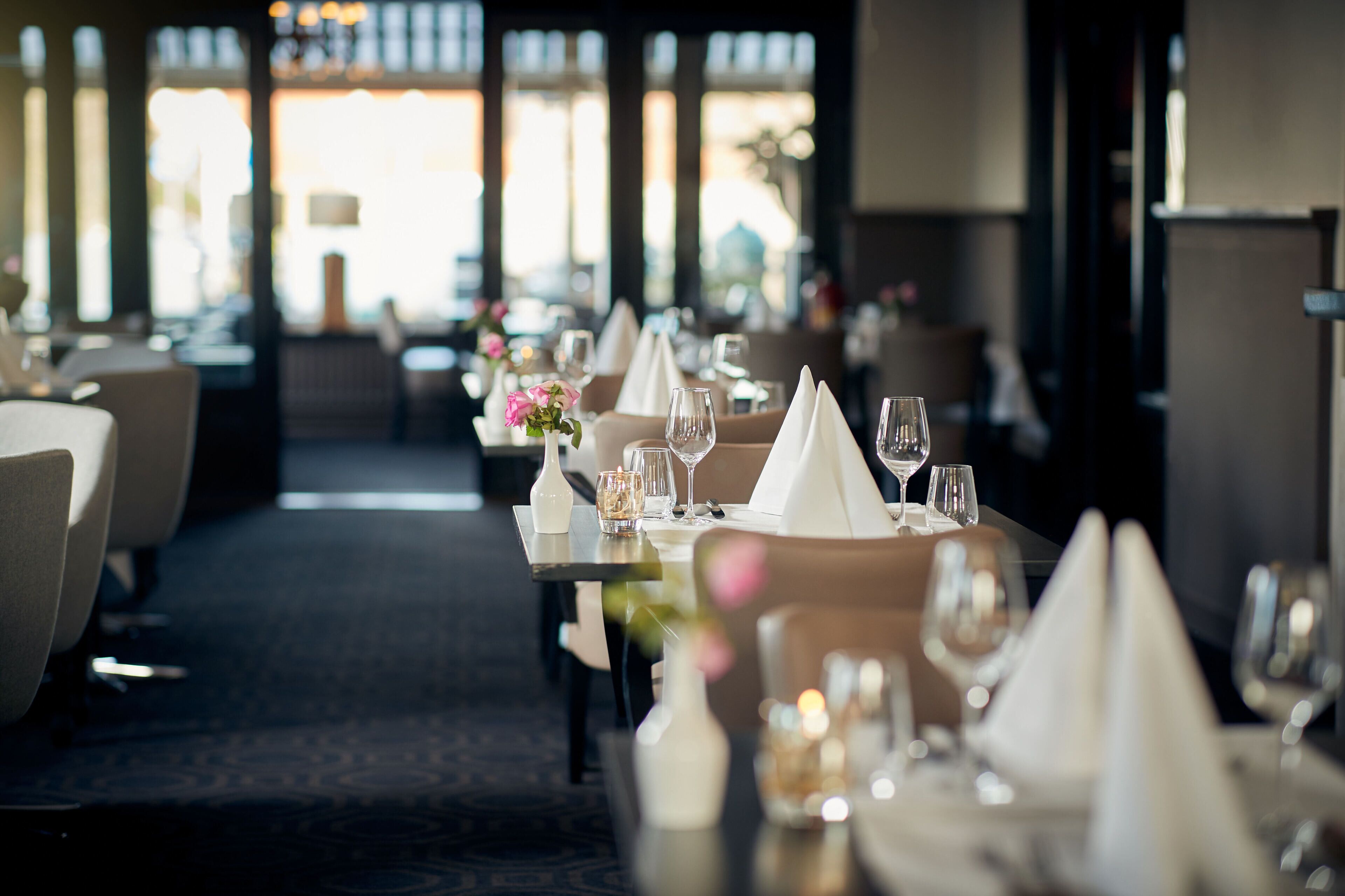 Ein Restaurant mit gedeckten Tischen, mit weißen Servietten, Weingläsern und kleinen Blumenvasen.