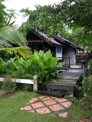 Una casa de madera tradicional con techo de paja, rodeada de abundante vegetación y una terraza de madera que conduce a la entrada.