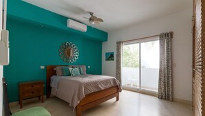 Une chambre à coucher comprenant un lit, une table de chevet, un banc, un ventilateur de plafond et une vue sur l’extérieur grâce à une porte-fenêtre coulissante.