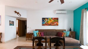 Un salon avec un canapé, une table basse et des fauteuils. Il y a un tableau au mur et un appareil de climatisation au-dessus du canapé.