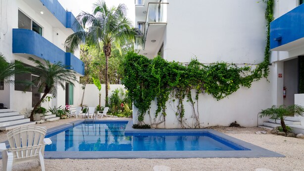Une piscine avec une chaise blanche, entourée d’une zone de gravier et d’un bâtiment aux accents bleus et agrémenté de verdure.