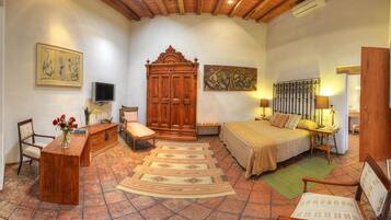 A bedroom with a wooden wardrobe, a bed with a metal headboard, a desk with a chair, a television, and a painting on the wall.