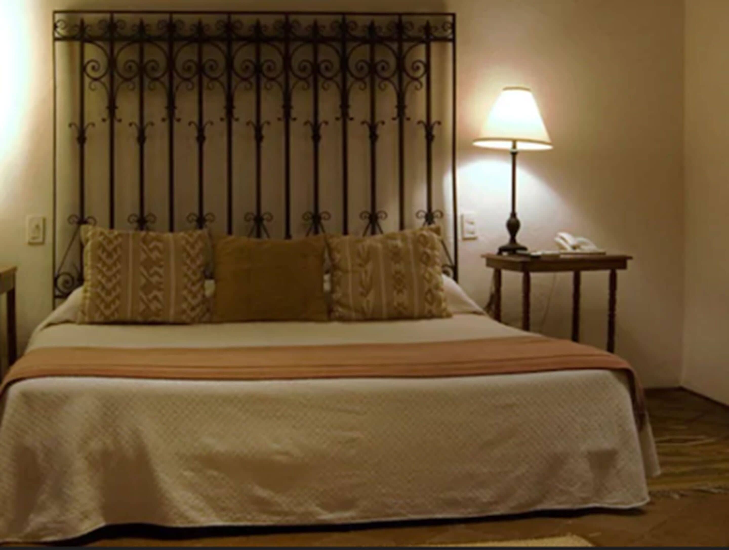 Una cama bien tendida con un cabecero estampado, una lámpara de noche y una mesita con un teléfono.