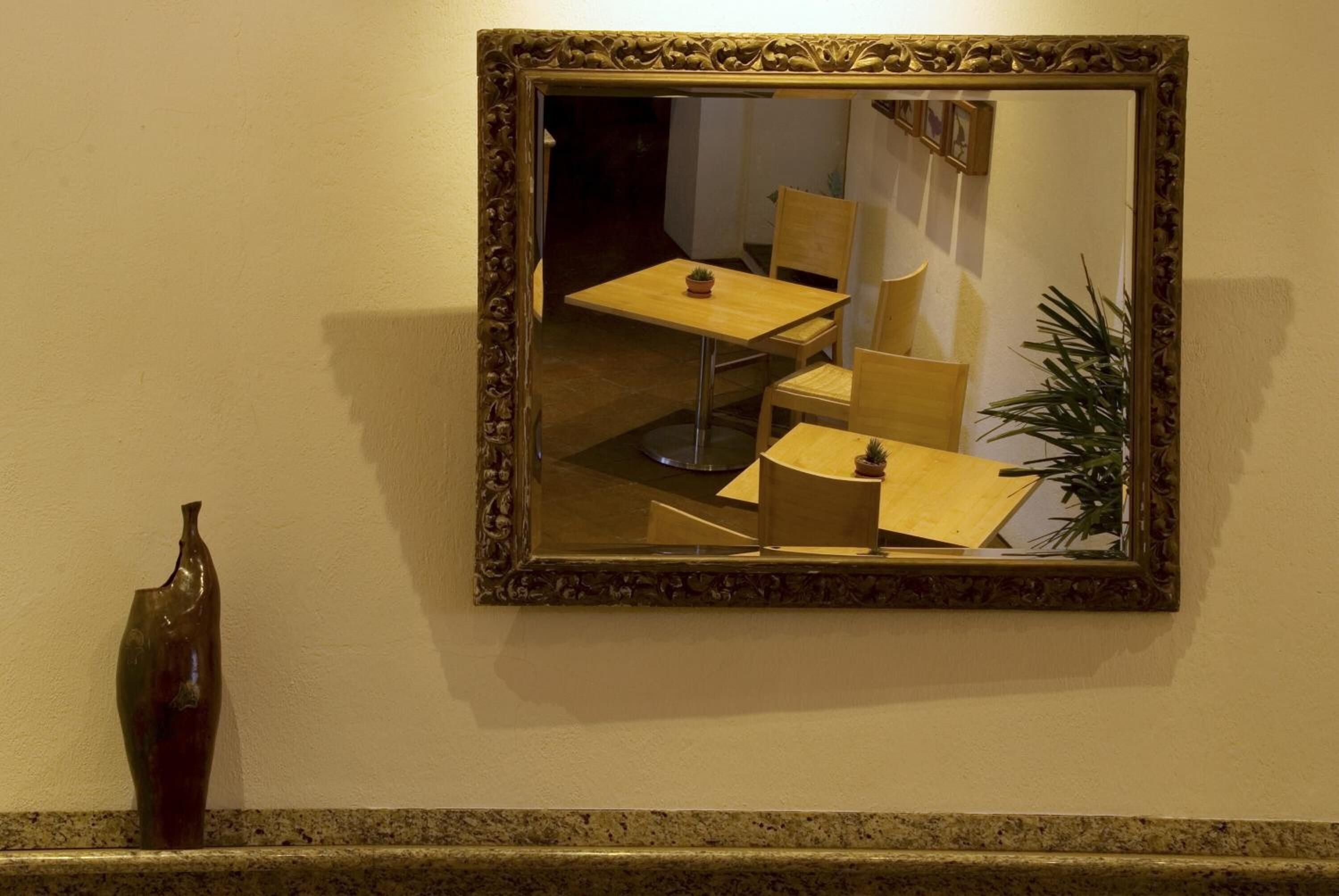 A mirror with a decorative frame reflecting a room with a table, chairs, and a plant.