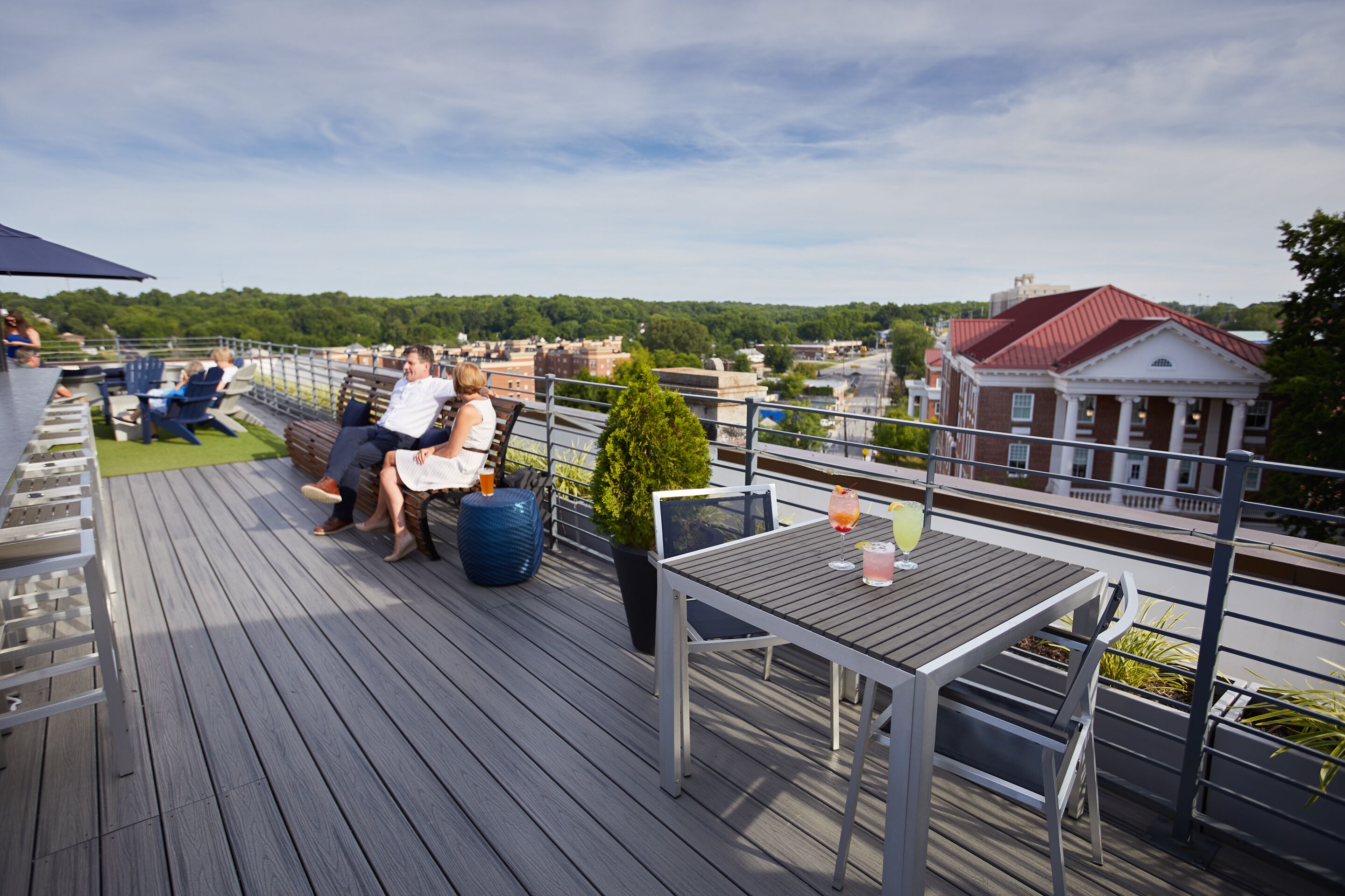 A rooftop deck with outdoor seating, a table with drinks, and a view of the cityscape.