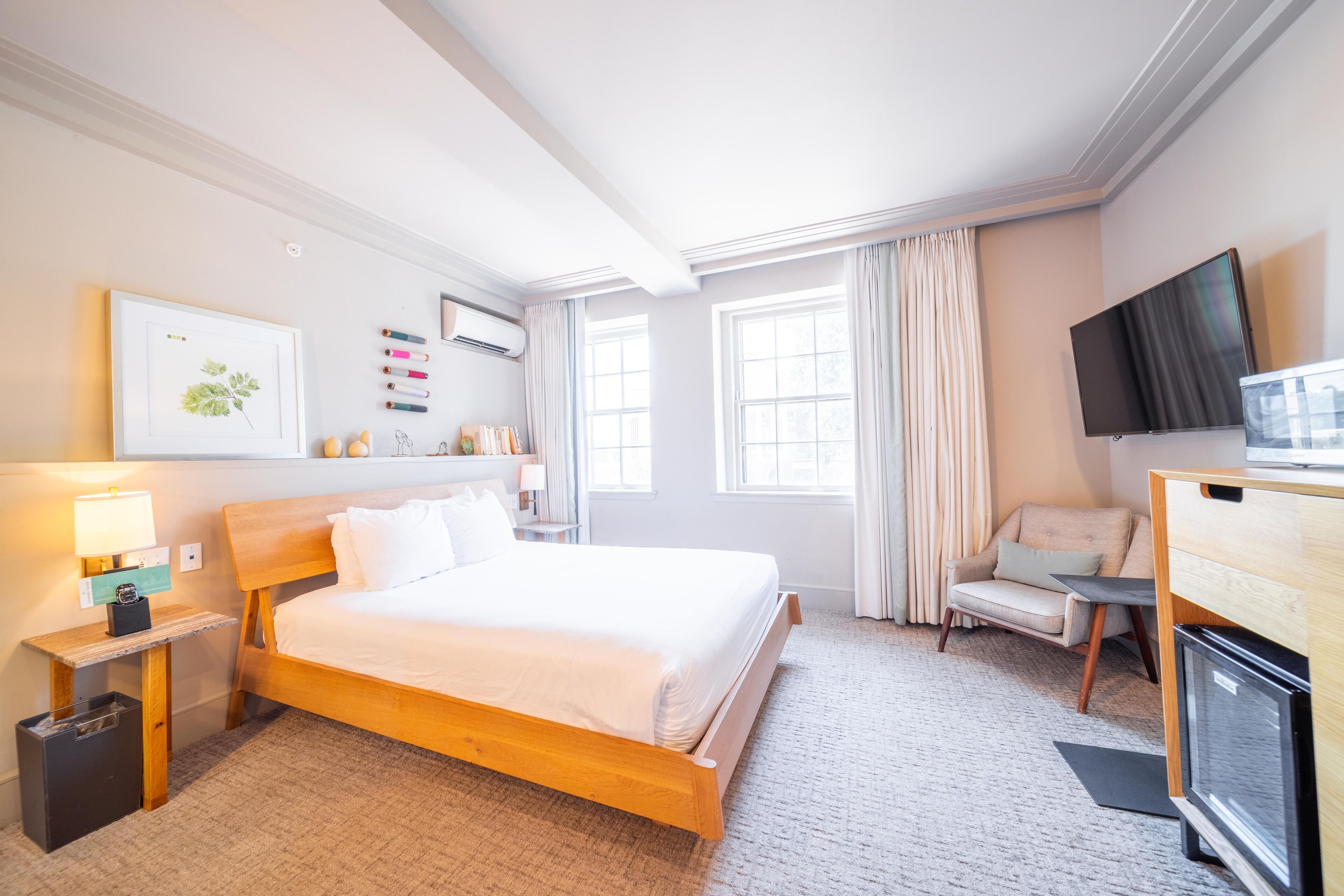 Habitación de hotel con una cama de madera, un escritorio con televisor, una silla y un ventanal con cortinas.