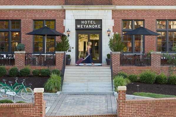 L’entrée d’un hôtel, avec une enseigne indiquant « HÔTEL WEYANOKE », des marches menant à la porte, et deux parapluies noirs placés de chaque côté.