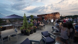 Een terras buiten met tafels en stoelen, potplanten en een bakstenen gebouw op de achtergrond.