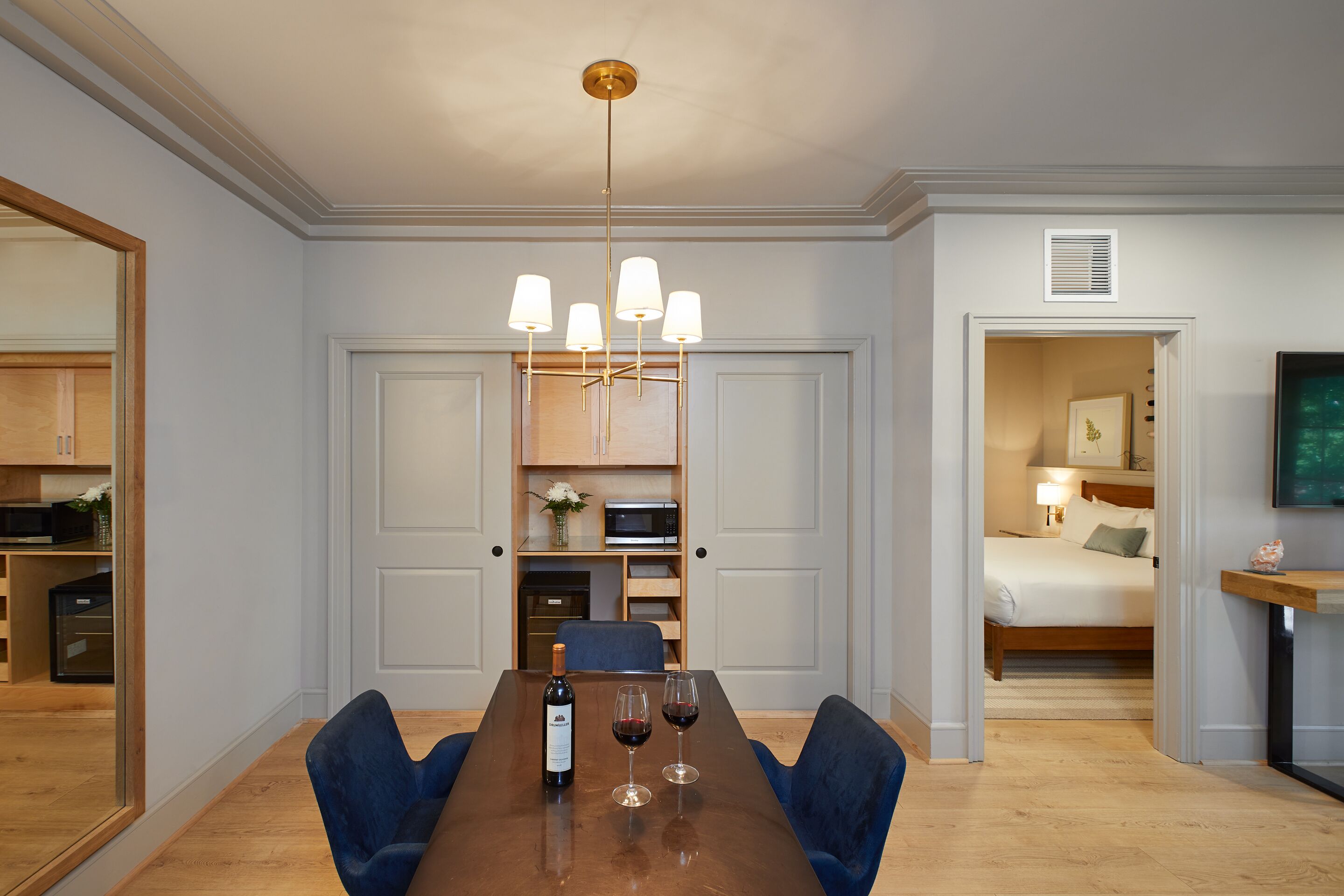 Una habitación de hotel moderna con una mesa de comedor de madera, sillas azules, un candelabro, una cama con una mesita de noche, y un televisor sobre un mueble de madera.