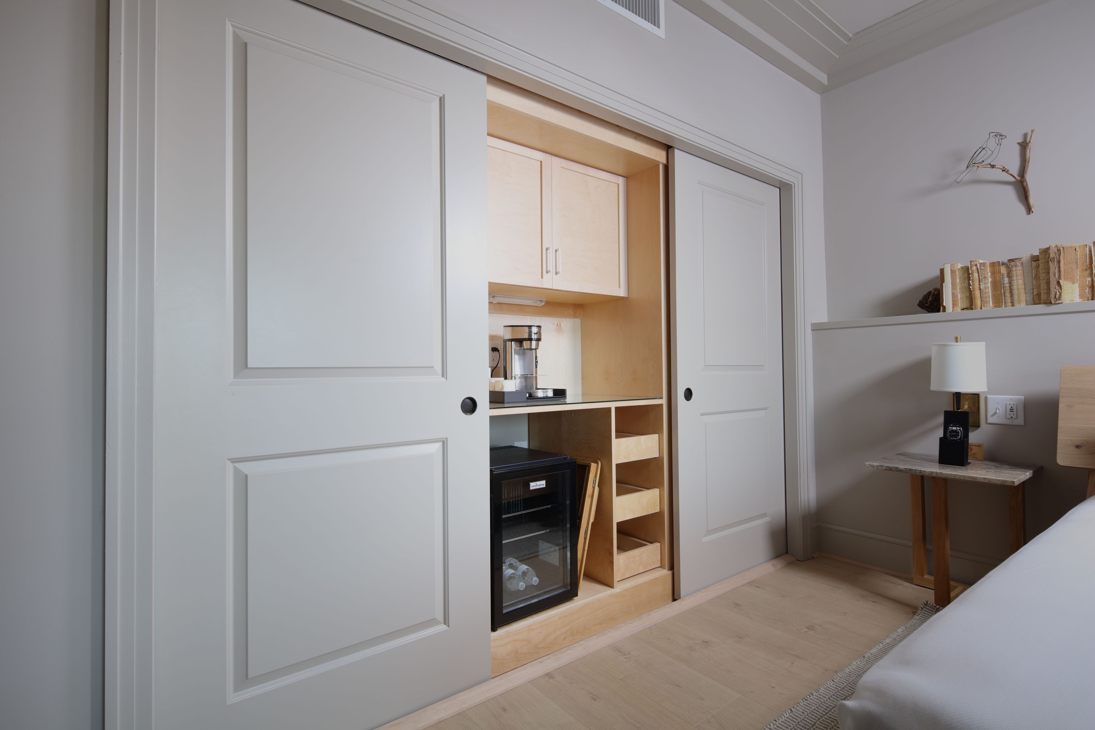 Una habitación con armario empotrado, una pequeña mesa de madera con lámpara y una estantería con libros.