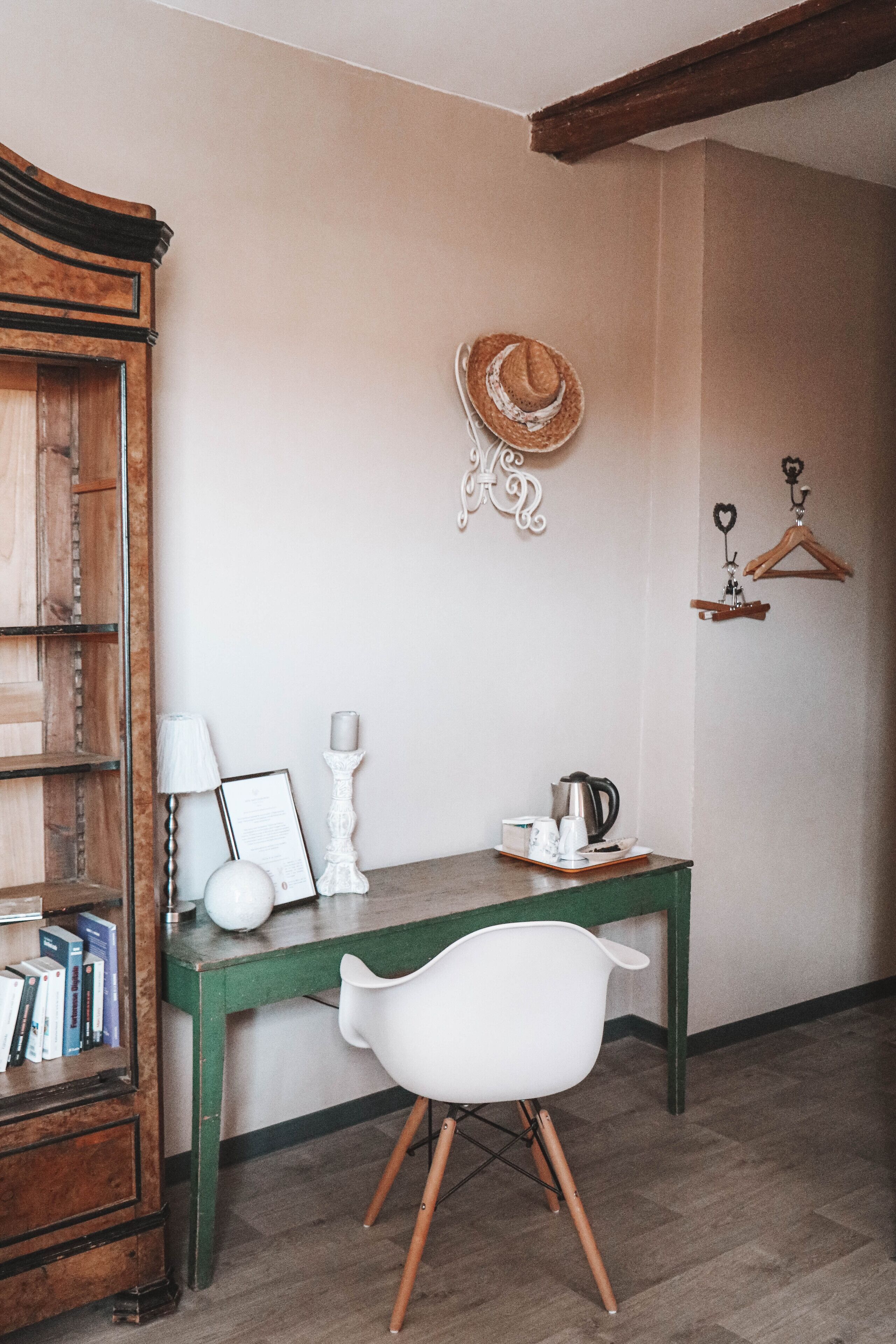 A room with a wooden desk, a white chair, a bookshelf, a hat on the wall, and a small shelf with items.
