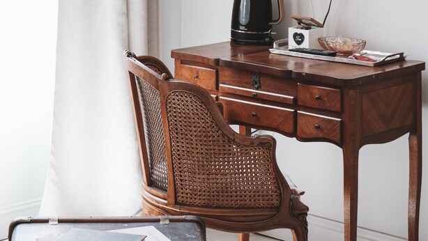 A room with a wooden desk, a television mounted on the wall, a wicker chair, and a suitcase. - Hotel Delga - Maison d'hôtes (Gaillac)