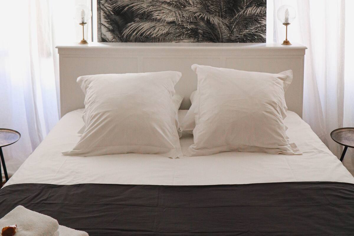 Une chambre avec un grand lit, des rideaux blancs et une décoration murale représentant des feuilles de palmier tropicales.
