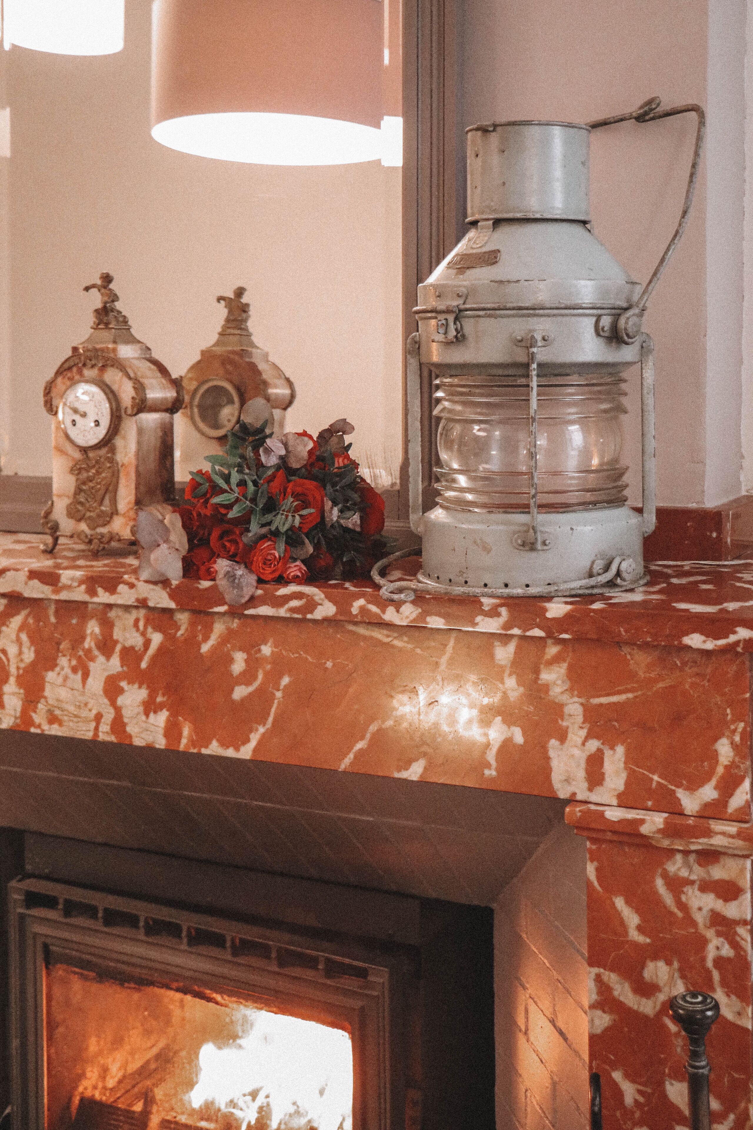 A fireplace with a mantel displaying a clock, a lantern, and a bouquet of flowers.