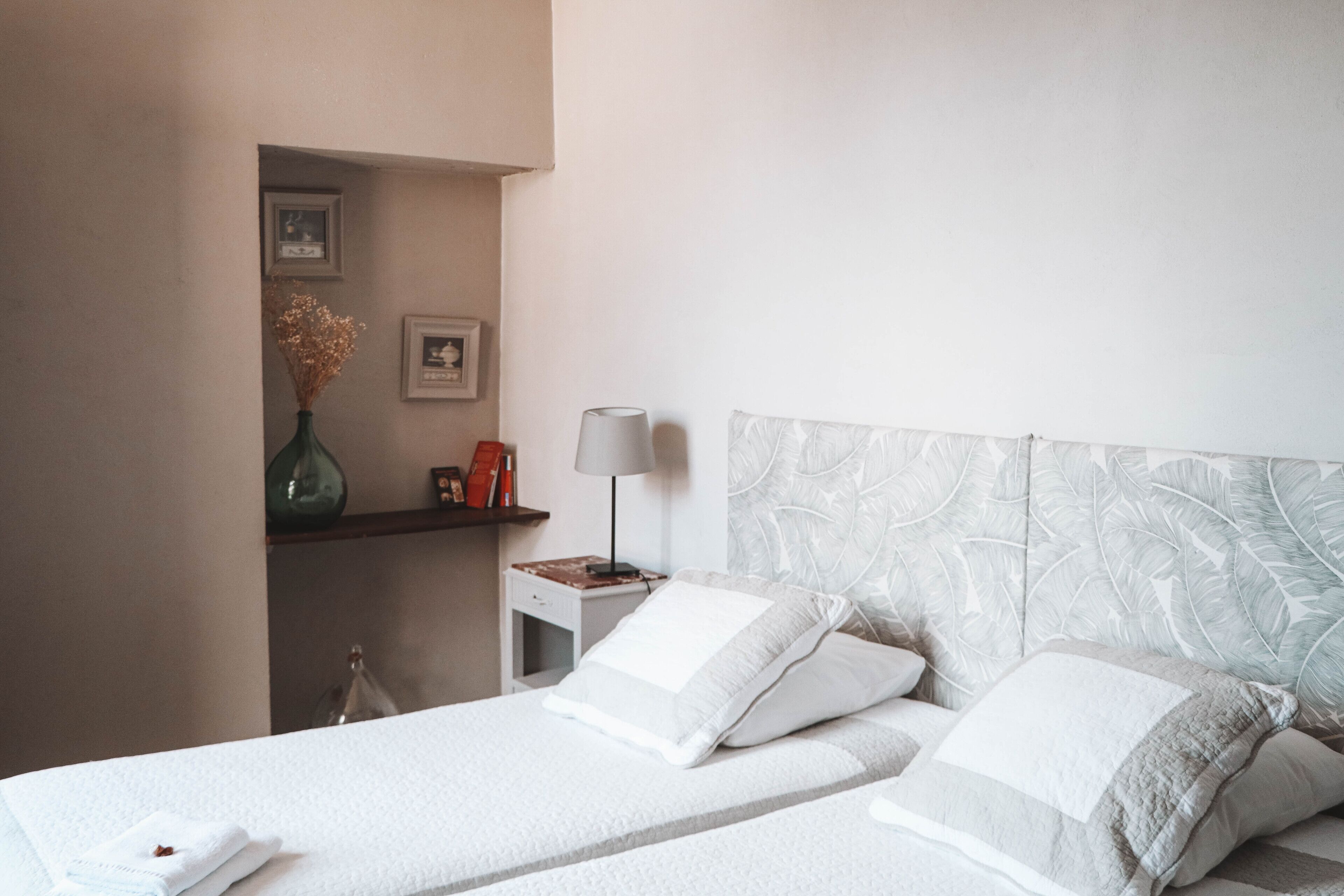 Ein Hotelzimmer mit einem Bett, einem Nachttisch, einer Lampe und einer Vase mit getrockneten Blumen.