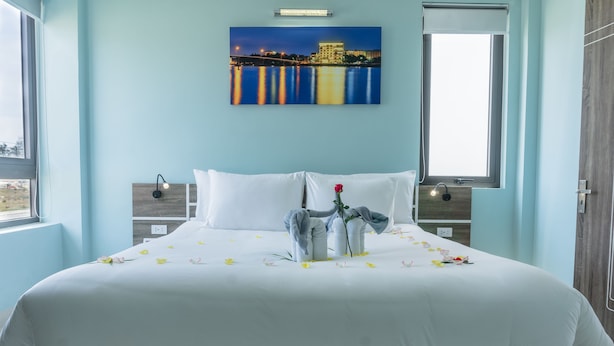 A neatly made bed with white linens, a single red rose, and scattered yellow flowers. A cityscape painting is above the bed. - Facetime Home (Da Nang)