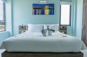 Een net opgemaakt bed met witte lakens, een enkele rode roos en verspreide gele bloemen. Boven het bed hangt een stadslandschap schilderij.