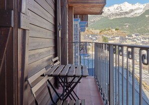 A wooden balcony with a table and benches, overlooking a river and a town with mountains in the background. - Aparthotel Tres Quiñones (Sallent de Gallego)