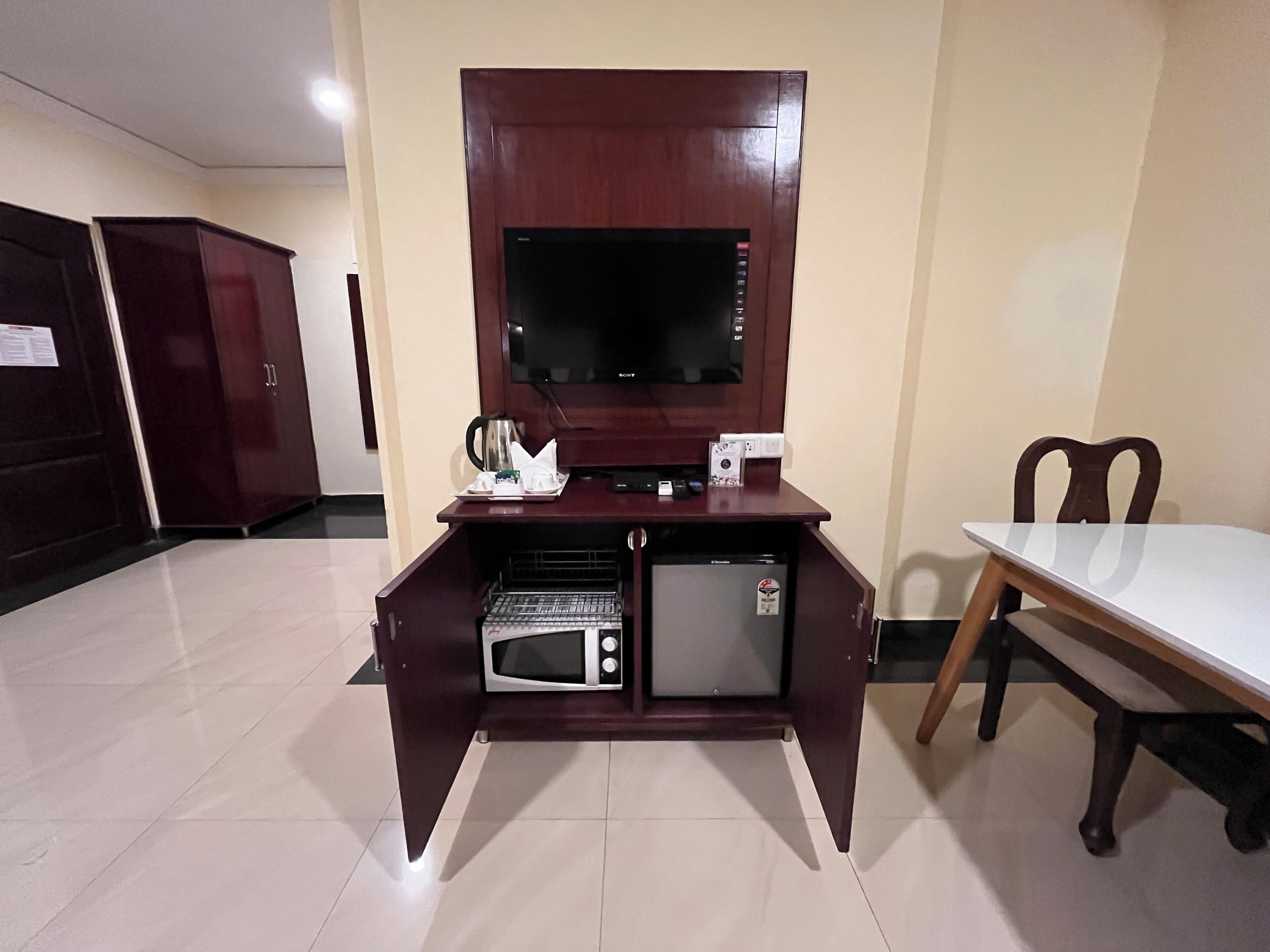 Il s’agit d’une pièce de taille modeste, dotée d’un bureau en bois, d’un micro-ondes encastré, d’un petit réfrigérateur et d’un téléviseur à écran plat. Une table et une chaise sont placées sur le côté droit.