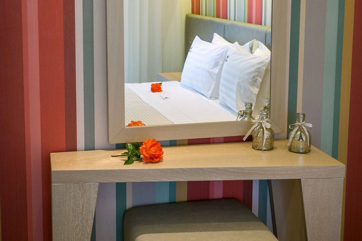 Una habitación de hotel con un espejo grande, una cama con sábanas blancas, una mesita de noche con botellas decorativas y una pared colorida a rayas.