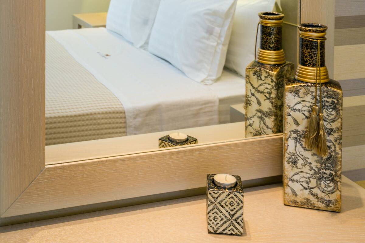 Una habitación de hotel con un espejo grande que refleja una cama bien hecha, una mesita de noche con jarrones decorativos y una mesita junto a la cama.