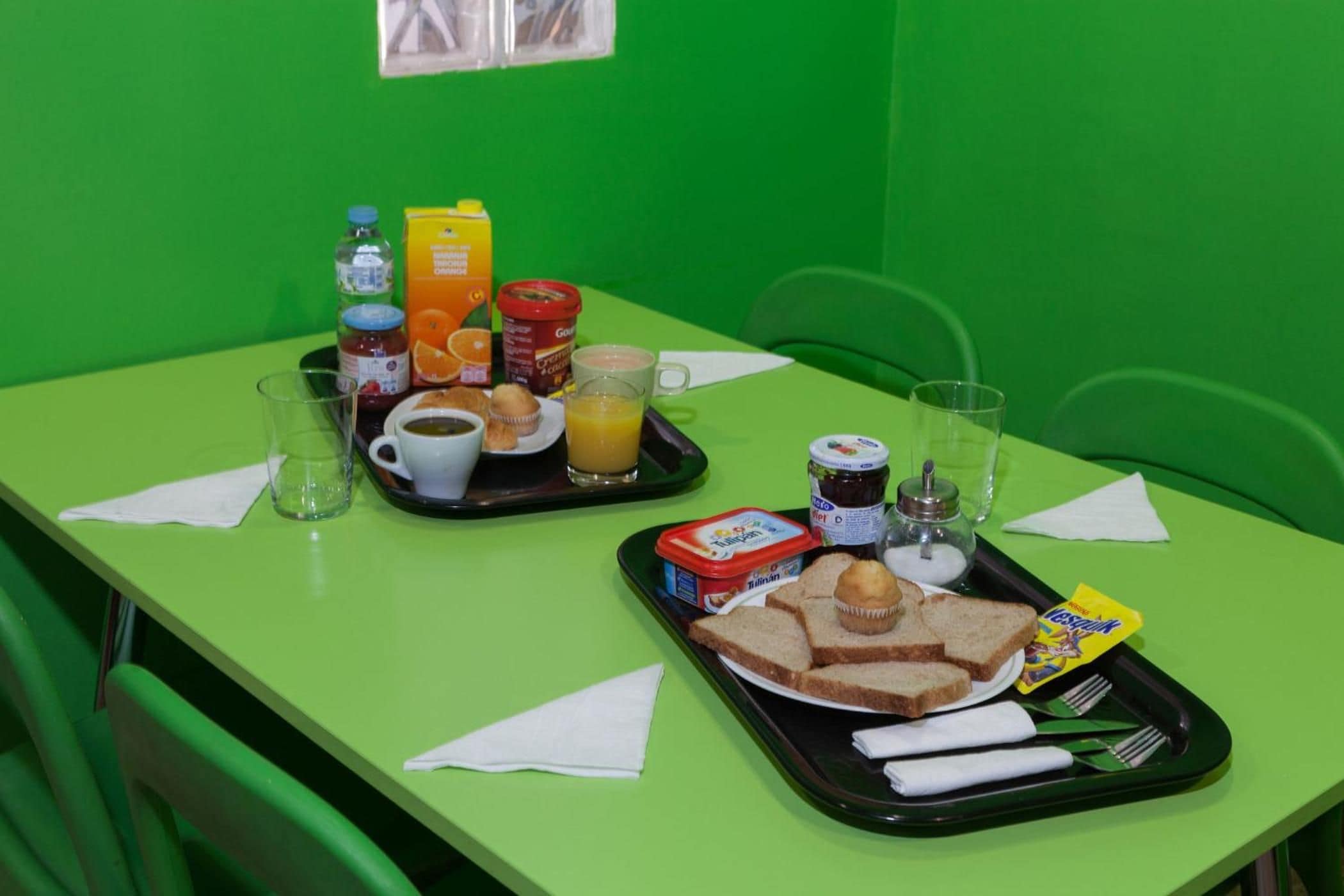 Bandeja de desayuno con tostadas, muffins, jugo y mermeladas sobre una mesa verde.