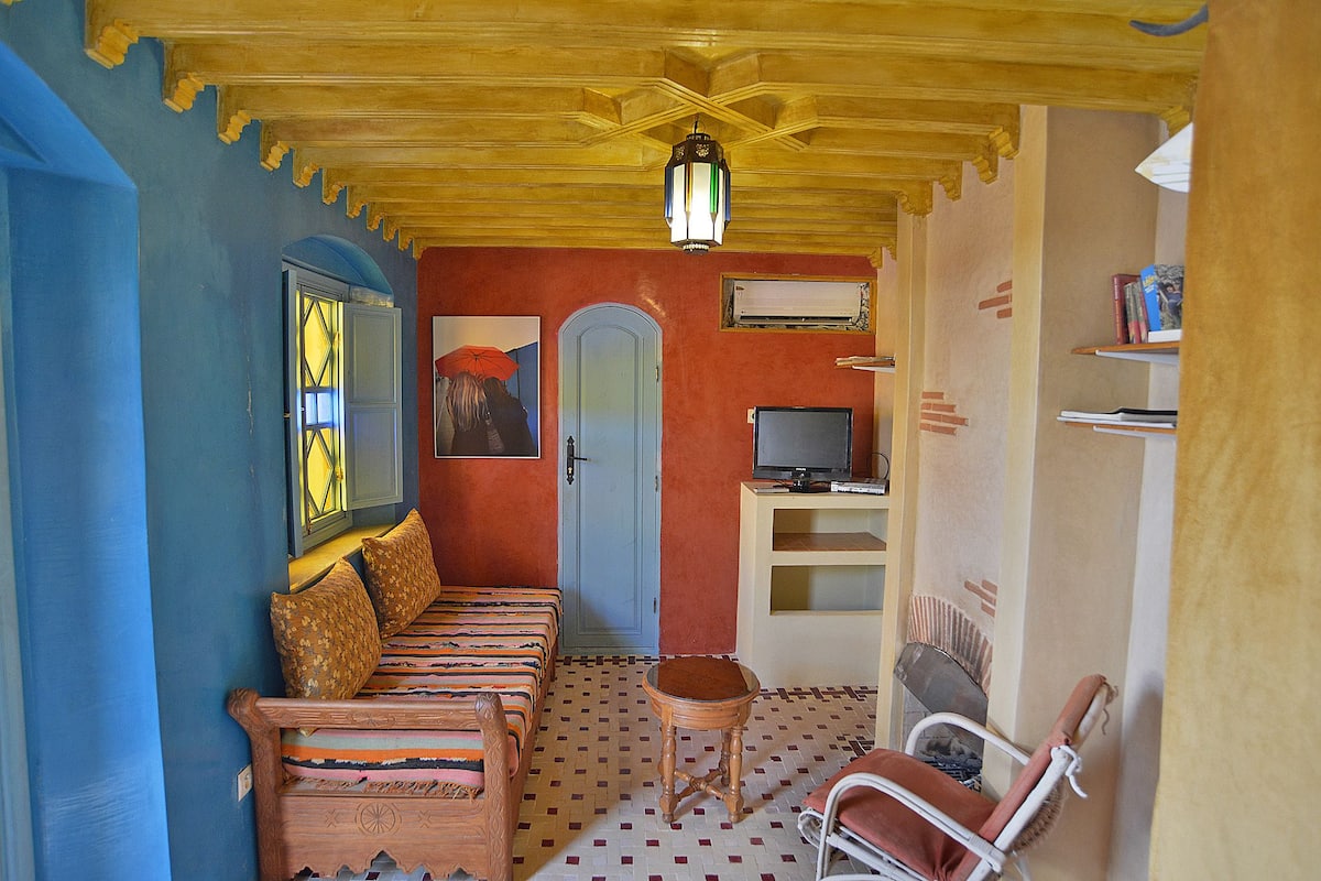 A room with a wooden ceiling, a sofa with patterned cushions, a television, and a small table with a chair.
