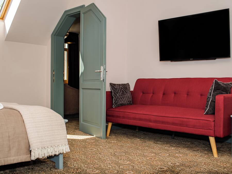 A room with a red sofa, a bed with a beige blanket, a patterned carpet, a television on the wall, and a door leading to another room.