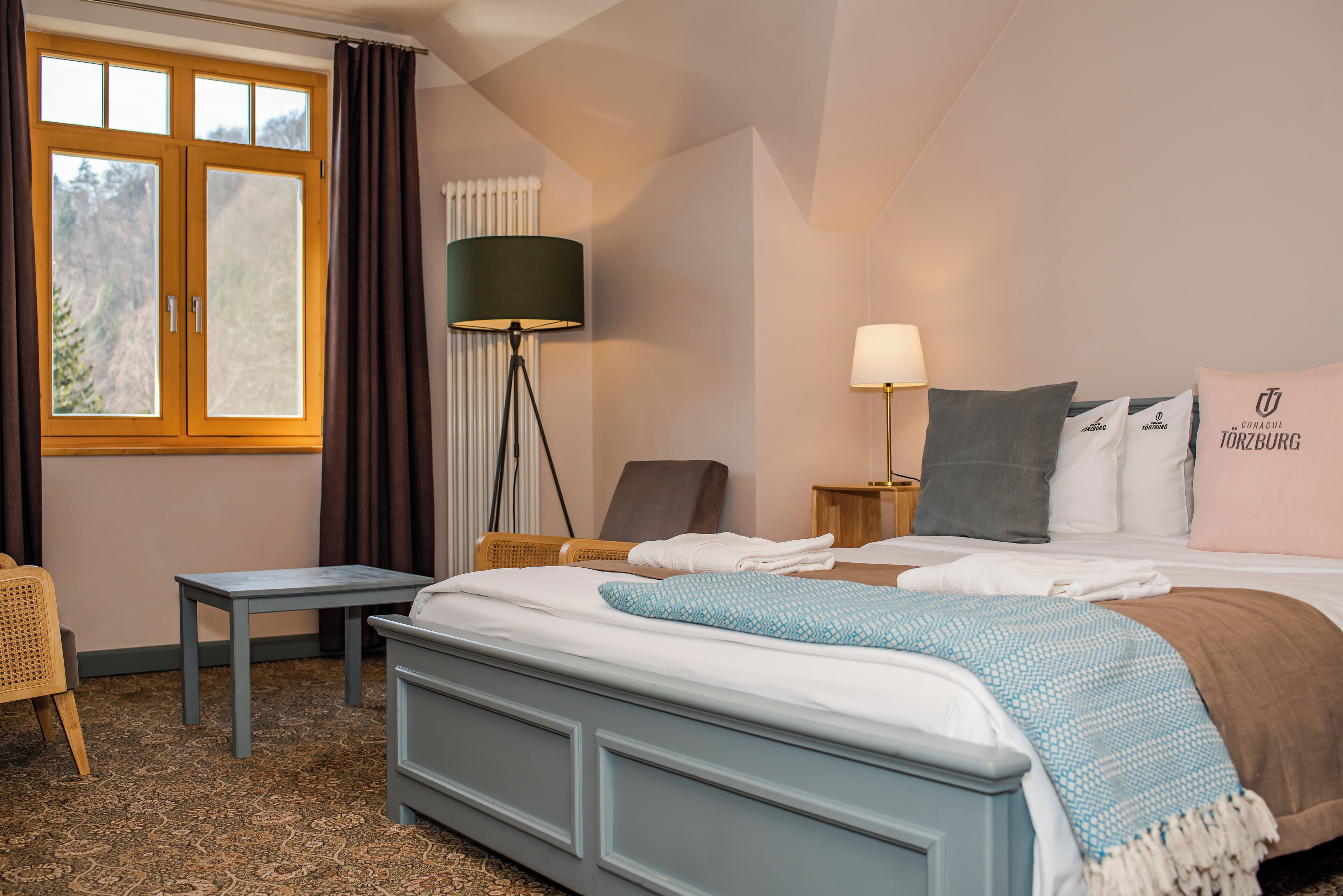 Una habitación de hotel con cama, mesita de noche, lámpara, silla y ventana con cortinas.