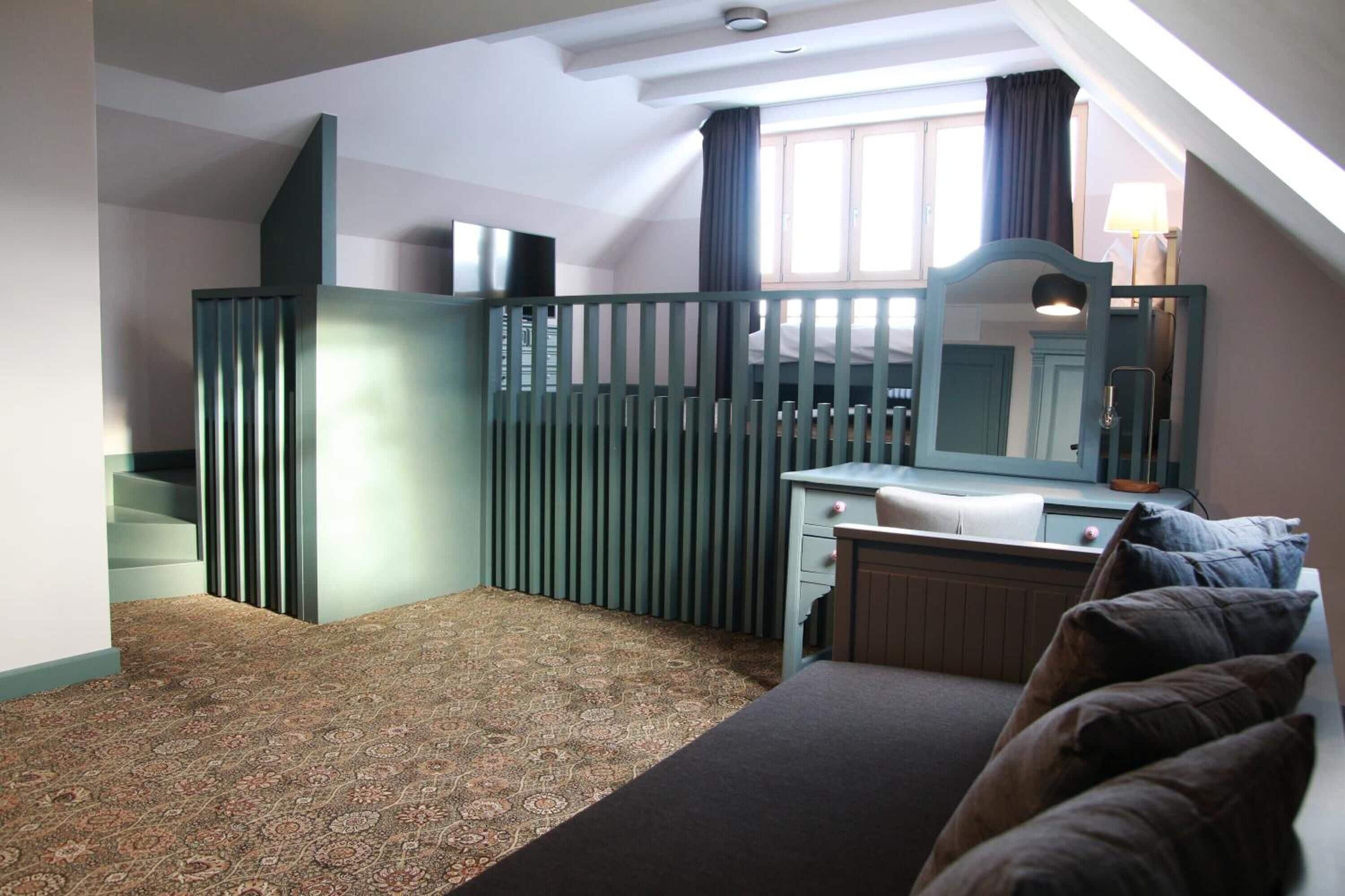 Ein Schlafzimmer mit Teppichmuster, einem Bett, einem Schminktisch und einem Spiegel, sowie einem Fenster mit Vorhängen.