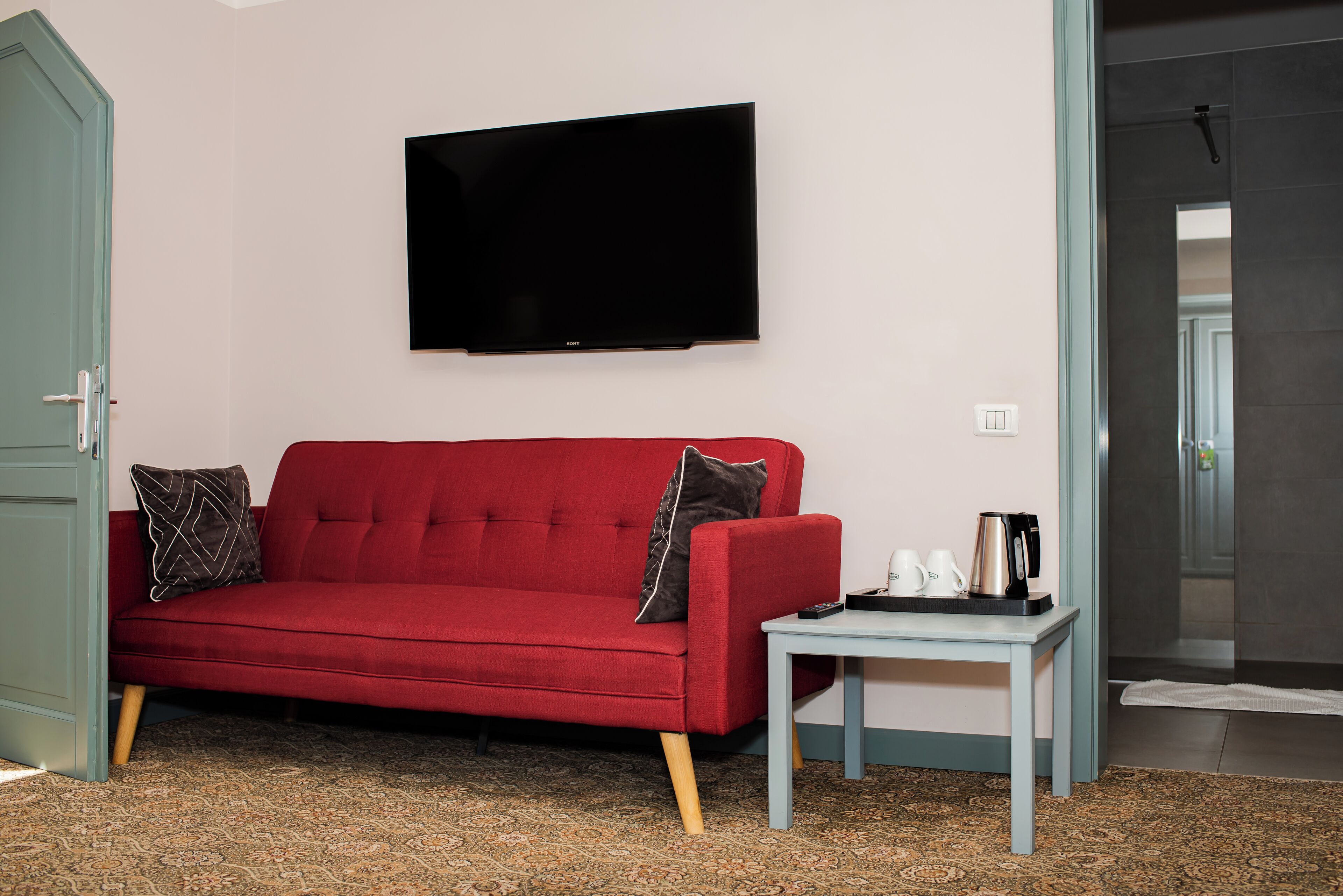 A red sofa with patterned cushions, a small table with a kettle and cups, and a flat-screen TV on the wall.