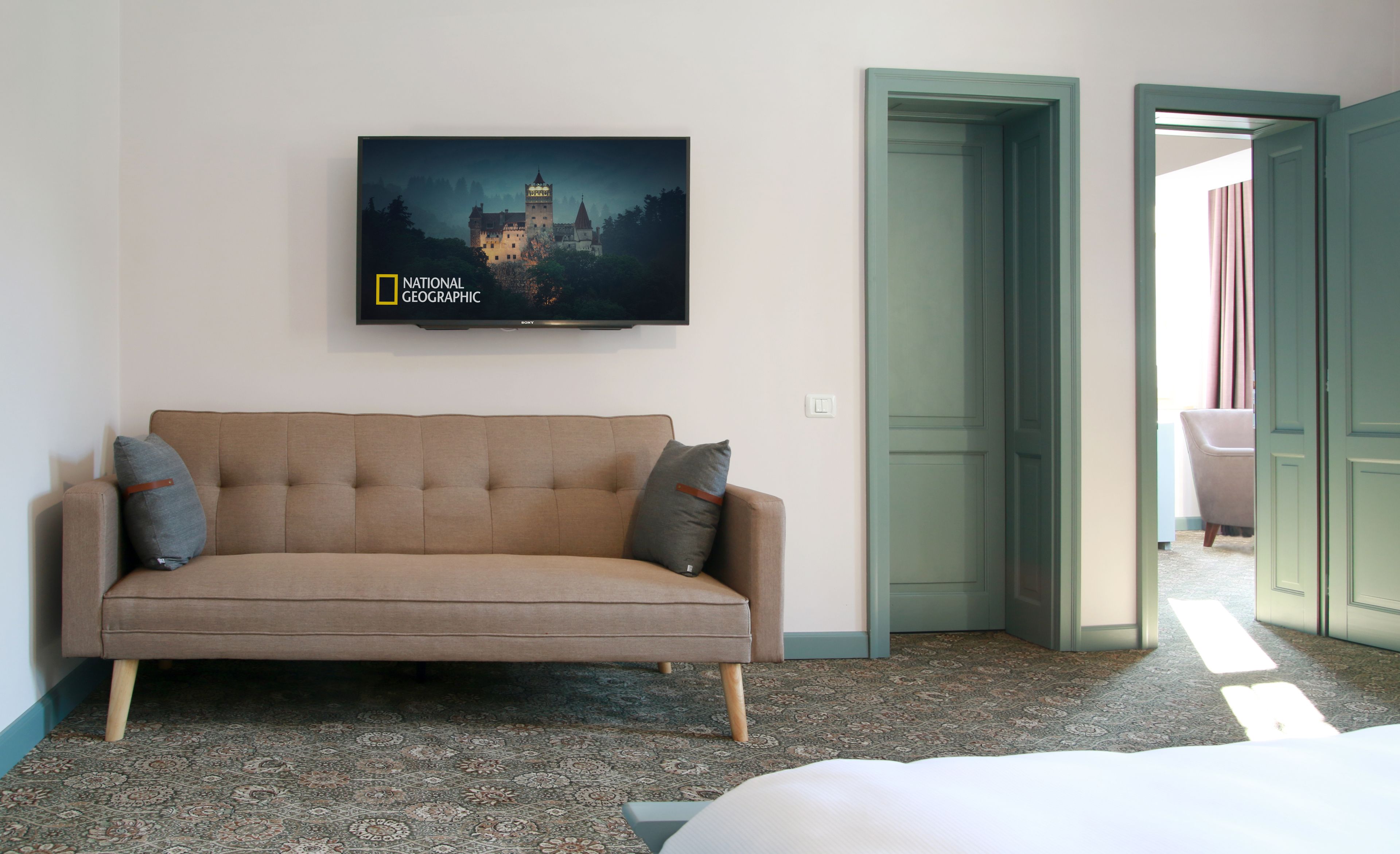 Habitación de hotel con sofá color beige, alfombra estampada, televisor mostrando una imagen de castillo y una puerta que da a otra habitación.