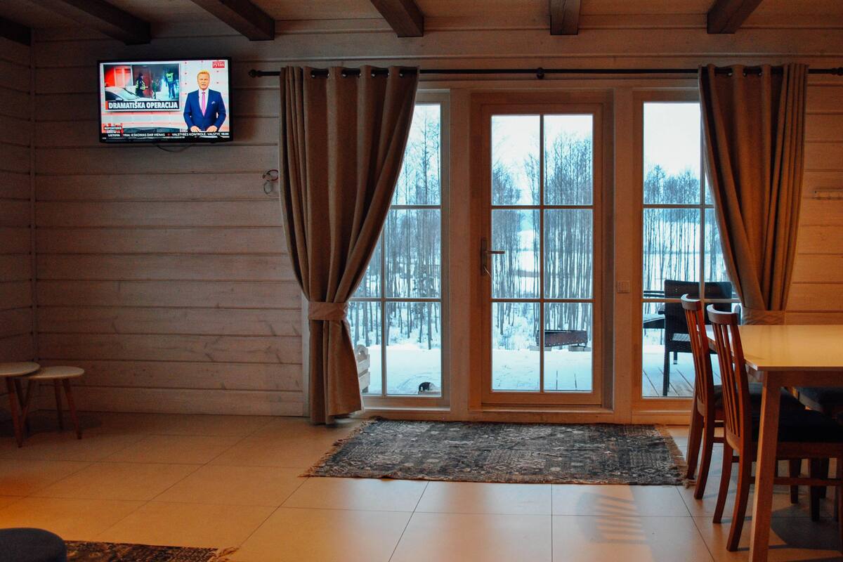 En inredning med träpaneler, en tv som visar ett nyhetsprogram, en matplats med ett bord och stolar samt utsikt över ett snötäckt landskap genom en glasdörr.