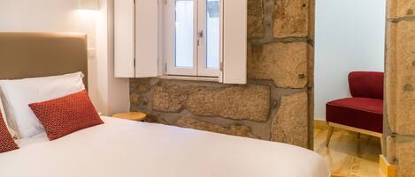 A bedroom with a stone wall, a bed with white and red bedding, a red chair, and a window with a view.