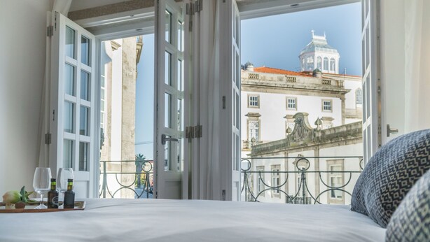 Habitació d'hotel amb un llit, una bandeja amb vasos i ampolles de vi, i una vista de cases antigues per finestres obertes.