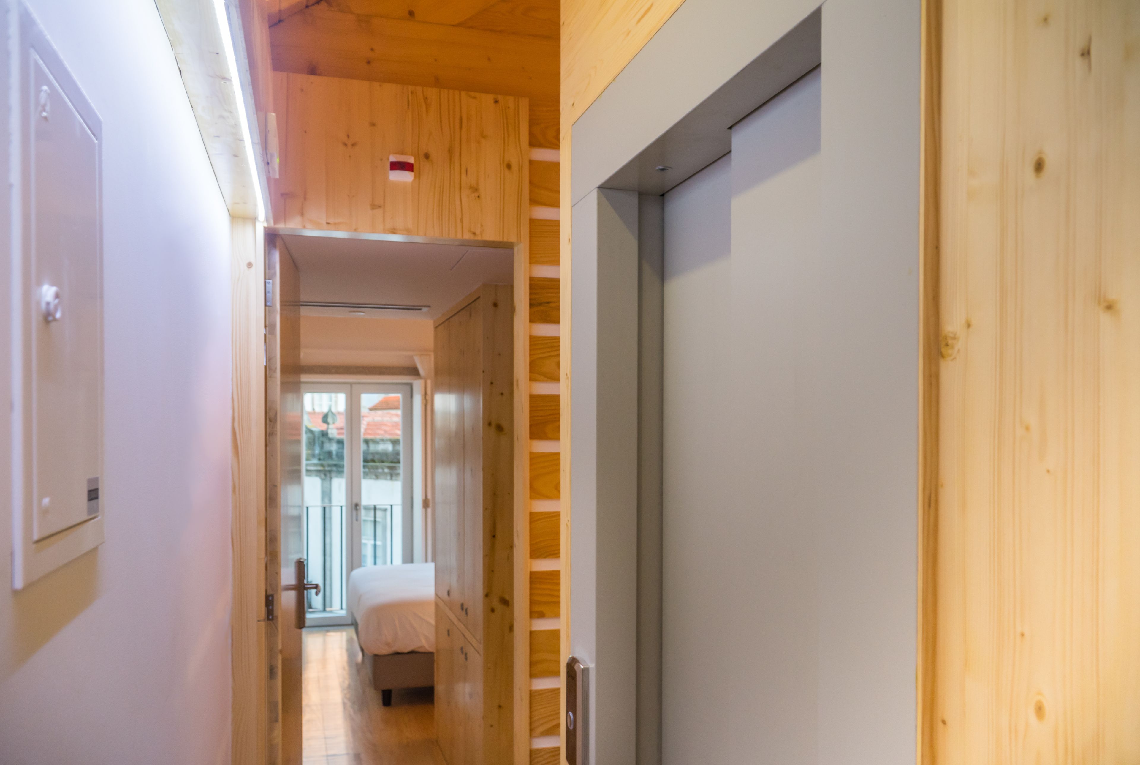 A narrow hallway with wooden walls and a door leading to a bedroom with a bed and a window.