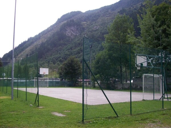 An outdoor basketball court with a netted enclosure, surrounded by grass and trees, with a mountainous backdrop. - Campodolcino Camping (Campodolcino)