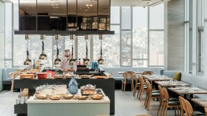 A modern dining area with a city view, a chef preparing food, and a variety of dishes displayed on the counter. - Novotel Ambassador Seoul Dongdaemun Hotels & Residences (Seoul)
