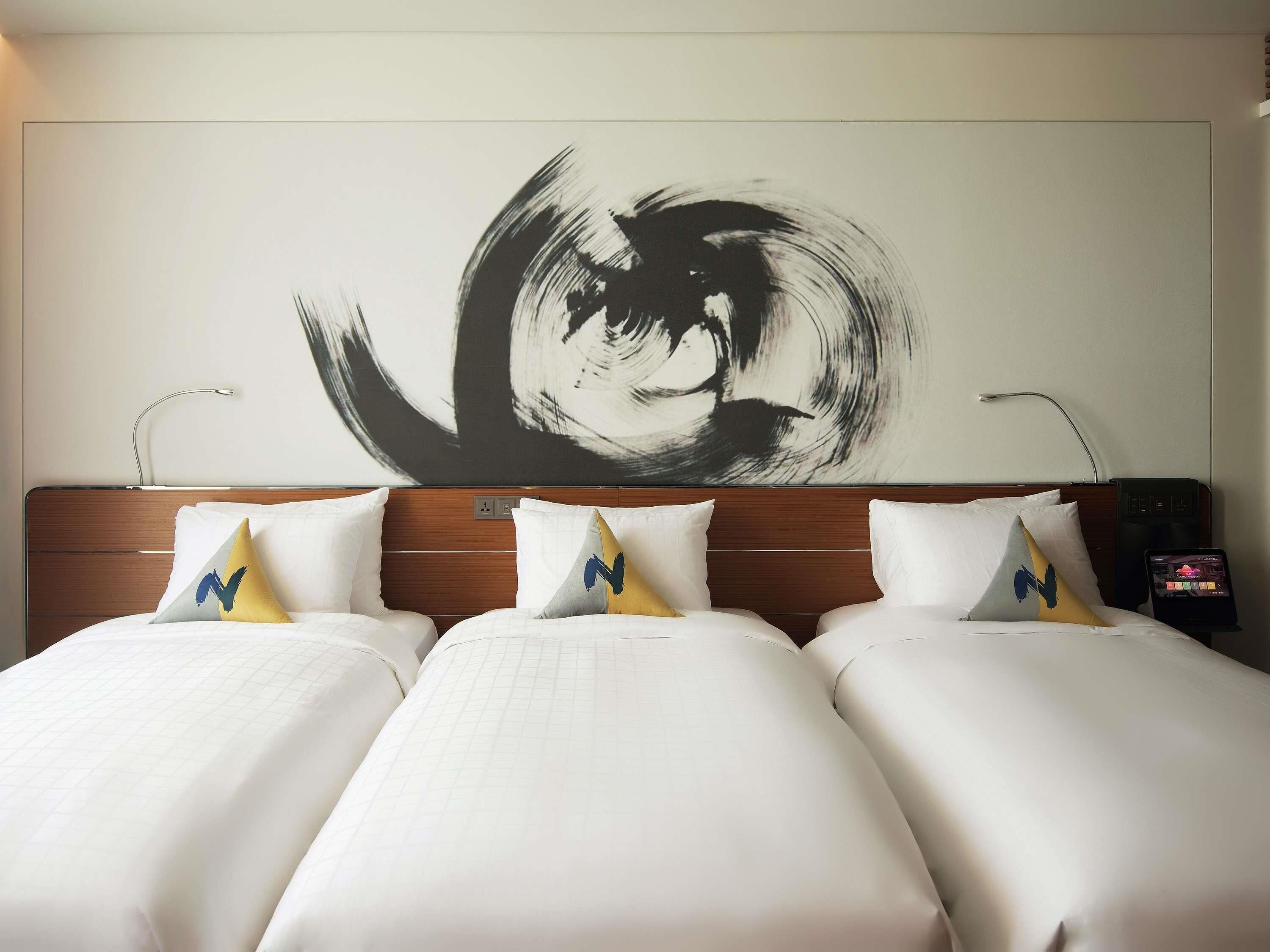 A hotel room with a large abstract painting on the wall, a wooden headboard, and a bed with white bedding and patterned pillows.