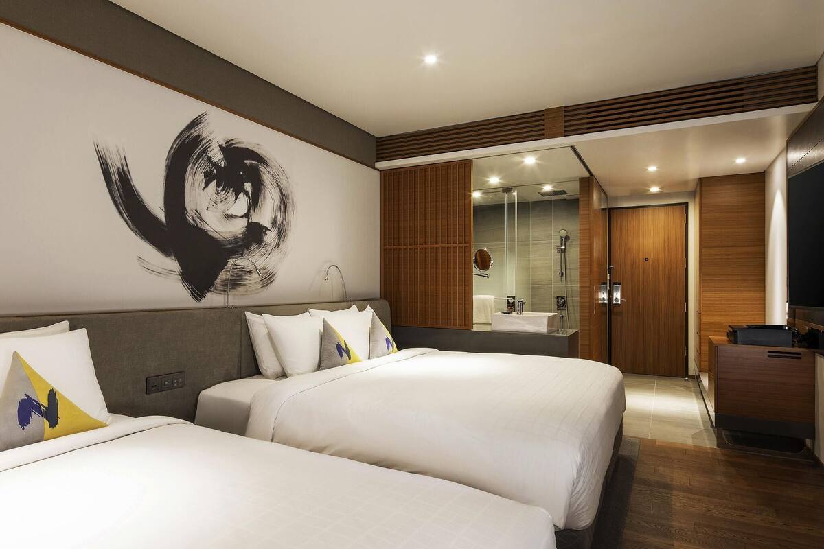 Habitación de hotel con dos camas, un cuadro grande en la pared, baño con puerta de vidrio y un armario de madera.