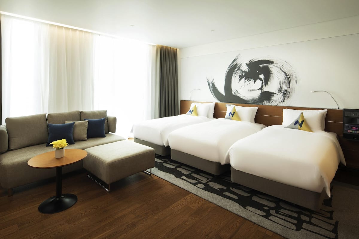 Une chambre d’hôtel avec un canapé, deux lits, une petite table ronde et un grand tableau abstrait accroché au mur.