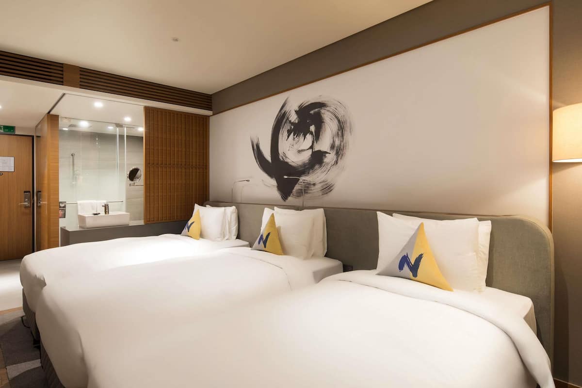 Une chambre d’hôtel avec deux lits, une grande peinture abstraite au mur et une salle de bain moderne dotée d’une douche en verre.