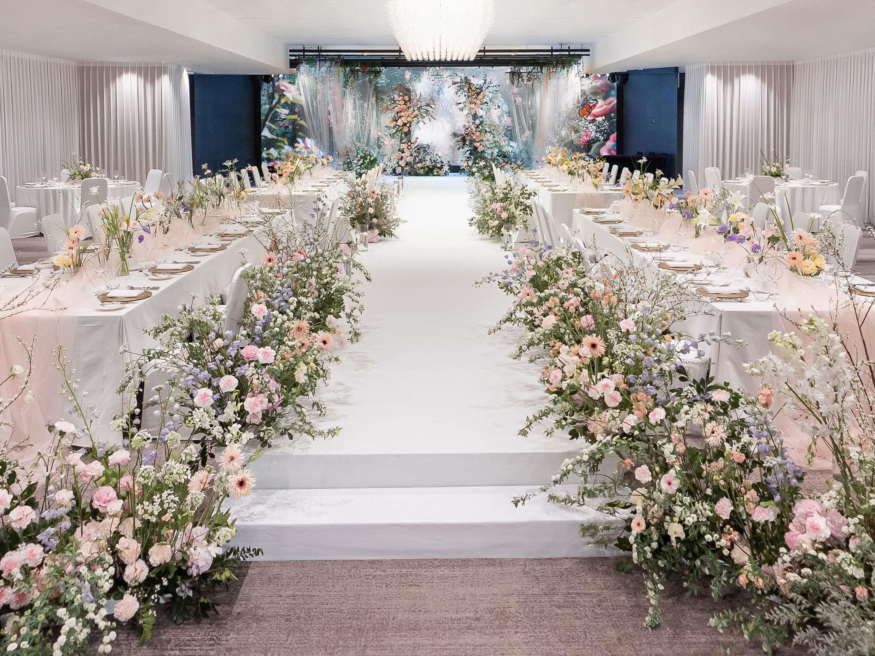 A banquet hall with a long table set for a formal event, flanked by floral arrangements.