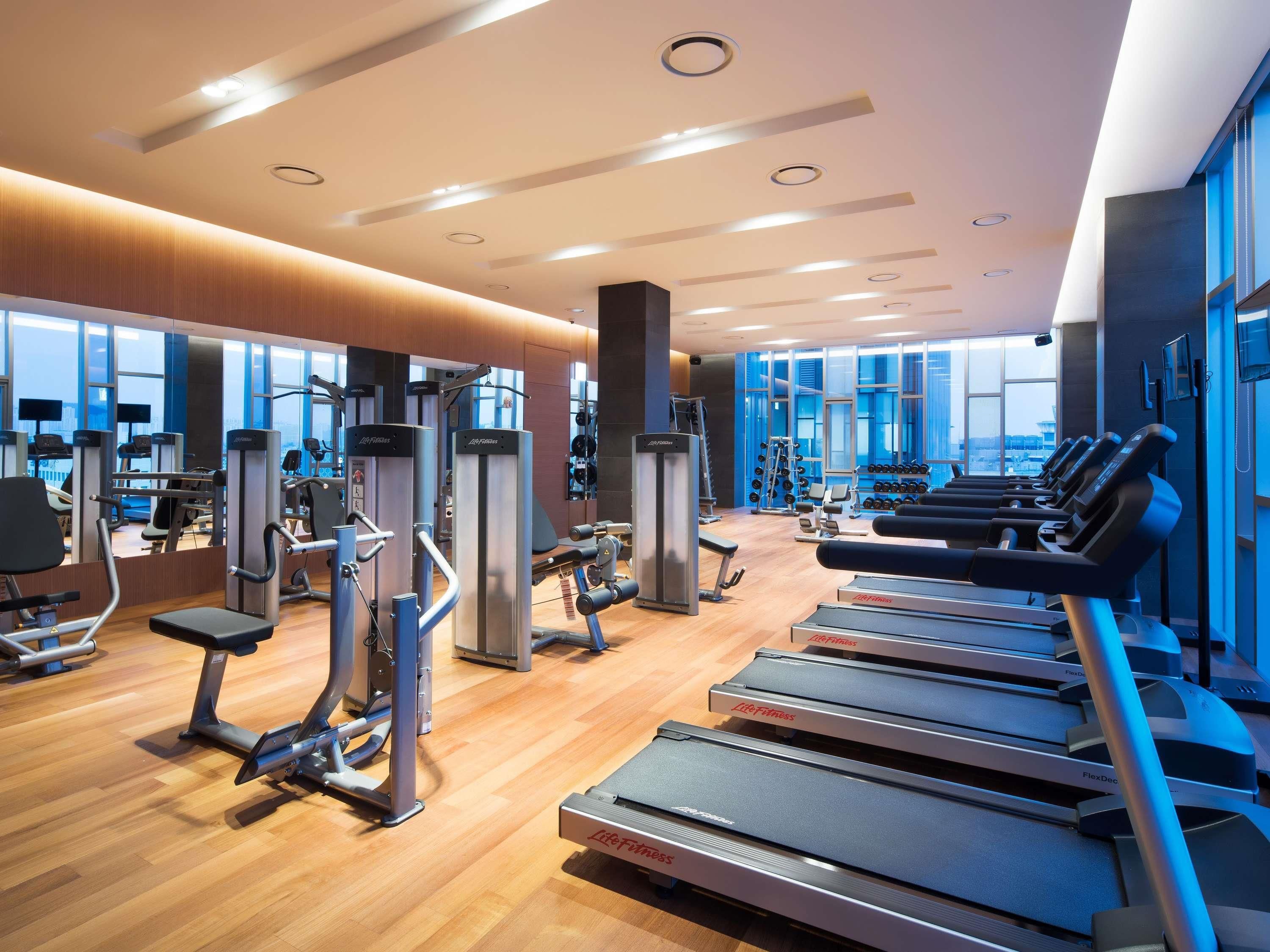A modern gym with cardio equipment, weight machines, and a large window providing a view of the city.