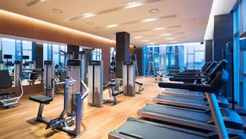 A modern gym with cardio equipment, weight machines, and a large window providing a view of the city.