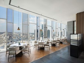 Una habitación de hotel moderna con ventanas amplias que ofrecen vistas a la ciudad, una zona de comedor con mesas y sillas redondas, y una kitchenette.