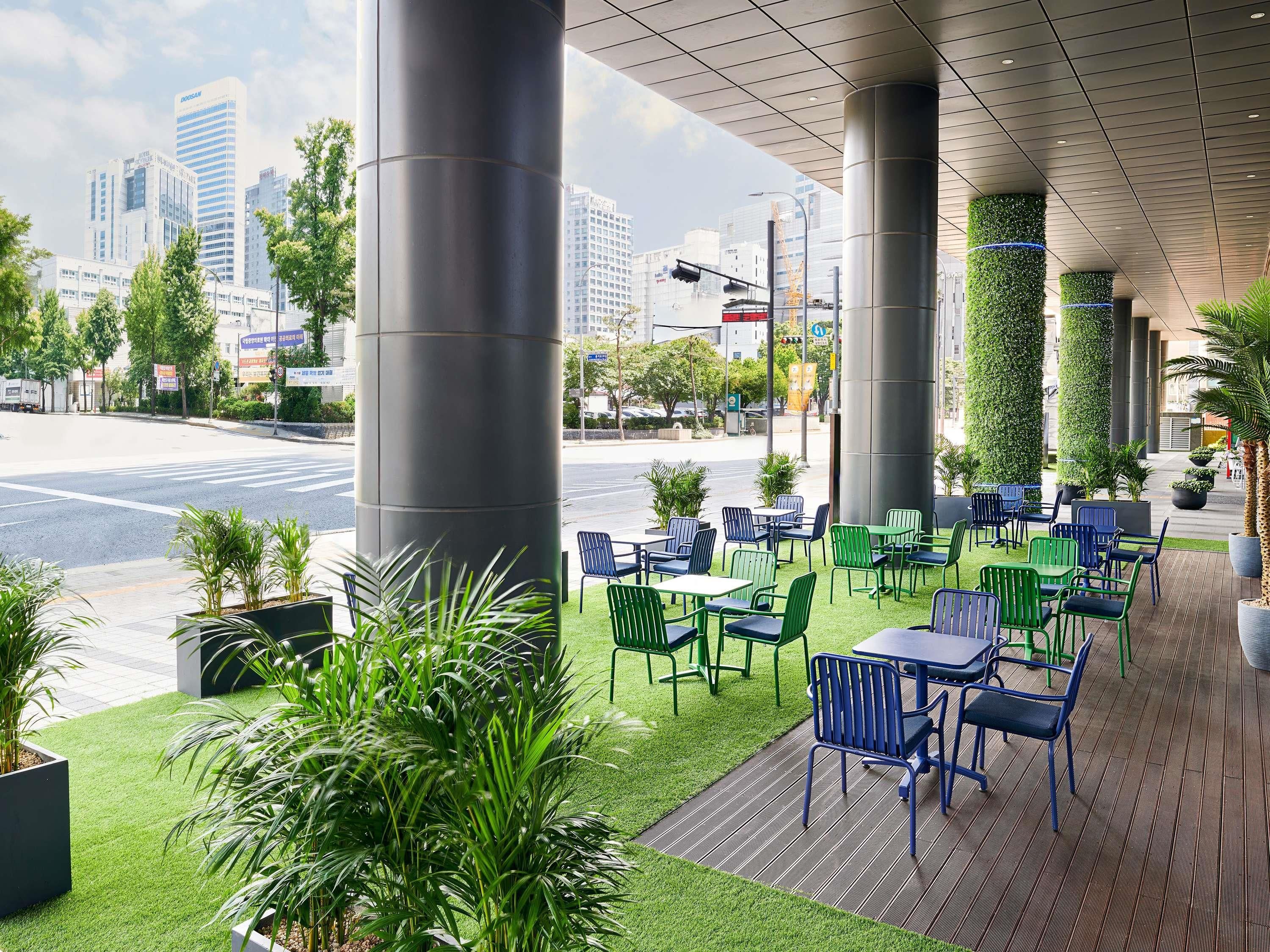Zona de estar al aire libre con sillas y mesas verdes y azules, rodeada de macetas y vistas a la ciudad.