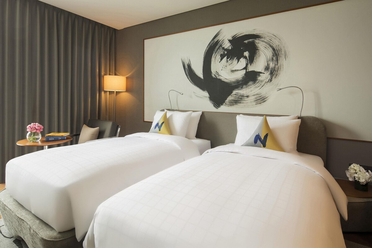A hotel room with two beds, a large abstract painting on the wall, a lamp, and a small table with a vase of flowers.