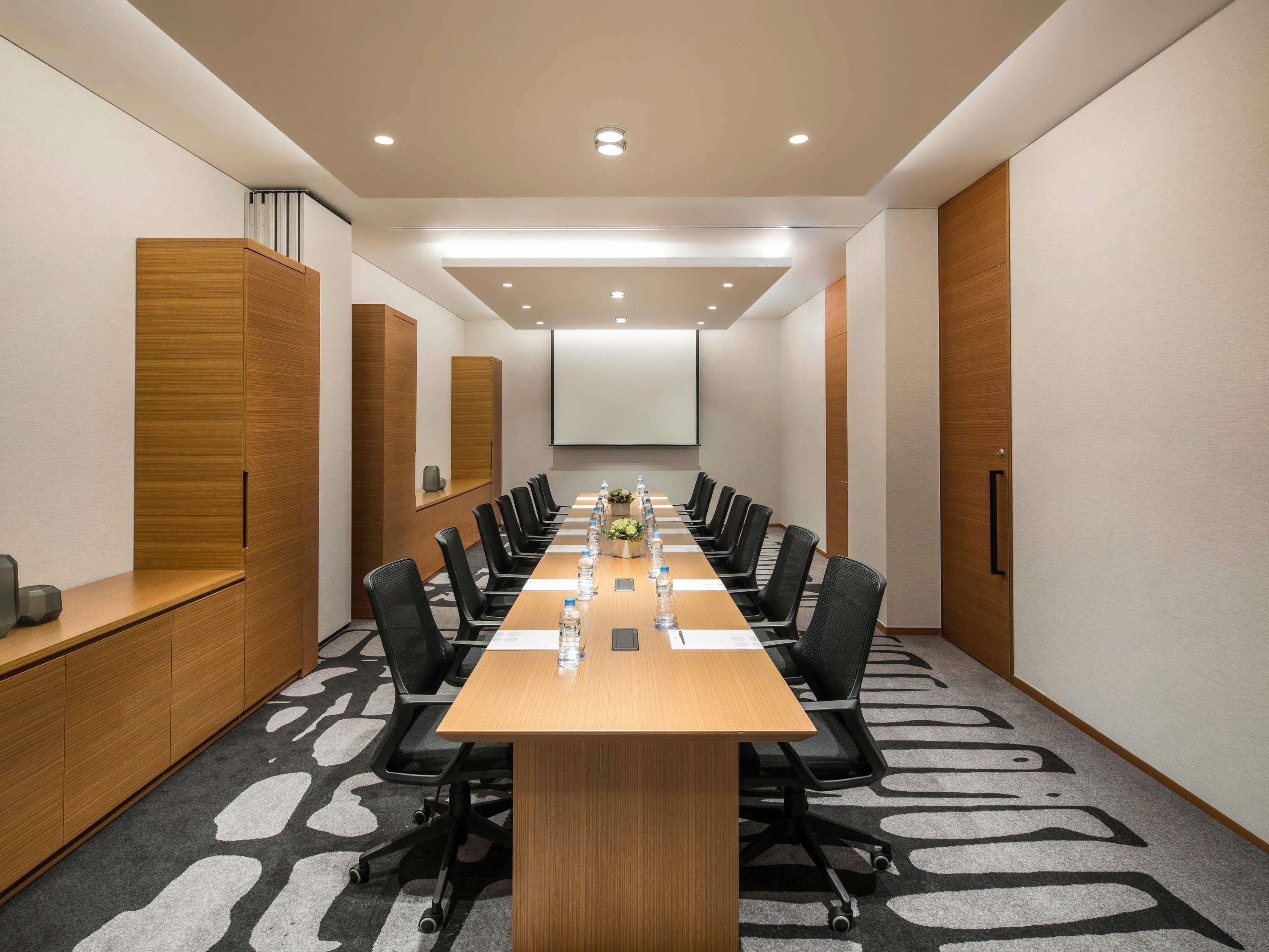 A conference room with a long table, black chairs, a projector screen, and a wooden cabinet.